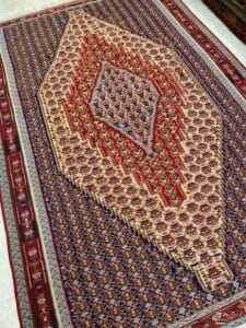 Azerbaijan Tappeti Milano Un colorato tappeto persiano con intricati motivi geometrici e floreali, prevalentemente nei toni del rosso, del blu e del beige, esposto su un pavimento bianco.