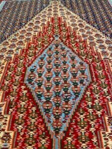 Azerbaijan Tappeti Milano Intricato tappeto persiano con motivi geometrici nei toni del rosso, del blu e del marrone, caratterizzato da una forma centrale a diamante. Un logo colorato con una lettera corsiva si trova nell'angolo in alto a destra.