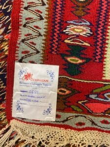 Azerbaijan Tappeti Milano Un tappeto colorato intrecciato con motivi geometrici e un'etichetta con informazioni dettagliate sul servizio di restauro e pulizia di Azerbaijani Restauro e Lavaggio Tappeti.