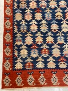 Azerbaijan Tappeti Milano Un tappeto con un motivo geometrico nei toni del blu, rosso e beige è esposto su un pavimento piastrellato chiaro. Il bordo presenta un motivo ripetuto e un logo a forma di foglia colorata è nell'angolo in alto a destra.