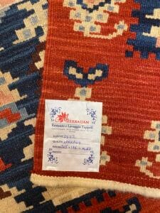 Azerbaijan Tappeti Milano Tappeto intrecciato colorato con motivi rossi, blu e beige parzialmente piegato. Un'etichetta mostra i dettagli di pulizia in italiano e menziona "Azerbaijan". È visibile un logo a farfalla multicolore.