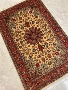 Azerbaijan Tappeti Milano Un tappeto rettangolare in stile persiano con intricati motivi floreali nei toni del rosso, del beige e del nero su uno sfondo chiaro, esposto su un pavimento piastrellato.