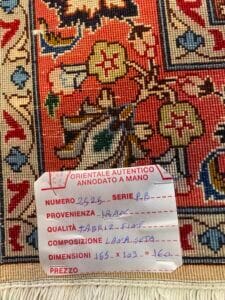 Azerbaijan Tappeti Milano Un primo piano di un tappeto a motivi con un'etichetta attaccata. L'etichetta elenca dettagli come l'origine del tappeto (Iran), la qualità, la composizione (lana e seta), le dimensioni e il prezzo.