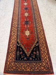 Azerbaijan Tappeti Milano Tappeto orientale lungo e rettangolare con centro rosso, intricati motivi geometrici, una sezione blu navy a un'estremità e un bordo dorato con motivi floreali ripetuti, esposto su un pavimento piastrellato chiaro. Nell'angolo in alto a destra è presente un logo colorato.