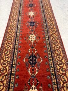 Azerbaijan Tappeti Milano Un tappeto rosso lungo e stretto con motivi geometrici e floreali in blu, beige e verde, bordato da disegni intricati. Nell'angolo in alto a destra è presente un logo astratto e colorato.