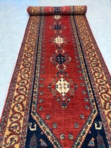 Azerbaijan Tappeti Milano Un tappeto rosso srotolato in stile persiano con intricati motivi geometrici e floreali nei colori crema, blu e oro, posto su un pavimento piastrellato. Nell'angolo in alto a destra si trova il logo di una foglia colorata.