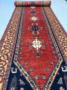 Azerbaijan Tappeti Milano Tappeto runner a fantasia rossa e blu navy con disegni geometrici e floreali è srotolato su un pavimento piastrellato; nell'angolo in alto a destra è visibile il logo di una foglia colorata.