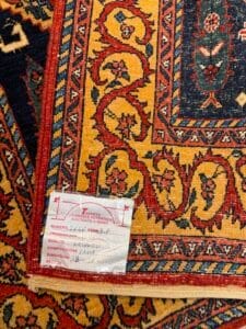 Azerbaijan Tappeti Milano Primo piano di un tappeto colorato con un'etichetta attaccata. L'etichetta contiene informazioni scritte a mano e stampate sull'origine, la qualità, la composizione e le dimensioni del tappeto. Il tappeto presenta intricati disegni in rosso, giallo e blu.