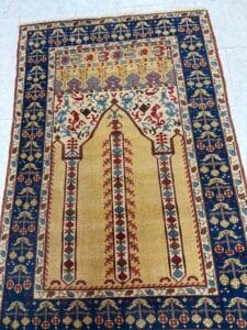 Azerbaijan Tappeti Milano Tappeto rettangolare con pannello centrale beige con motivi simmetrici rossi, blu e marroni, circondato da un bordo blu ornato con disegni floreali. La parte superiore presenta vivaci motivi intricati e un logo a farfalla multicolore.