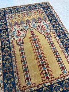 Azerbaijan Tappeti Milano Un tappeto colorato con un disegno centrale di mihrab beige con intricati motivi floreali e un bordo blu navy, esposto su un pavimento piastrellato chiaro. Nell'angolo compare il logo di una piccola foglia multicolore.