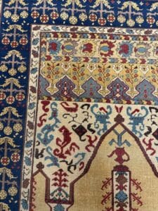 Azerbaijan Tappeti Milano Un tappeto colorato con intricati motivi geometrici e floreali nei toni del blu, rosso, giallo e crema. Una foglia artificiale multicolore si trova nell'angolo vicino al bordo superiore destro.