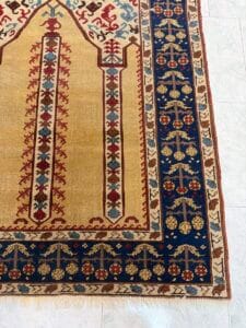 Azerbaijan Tappeti Milano Angolo di un tappeto decorativo con un motivo intricato, principalmente oro, blu e rosso. Nell'angolo in alto a destra compare il logo di una farfalla colorata, posizionata su un pavimento piastrellato chiaro.