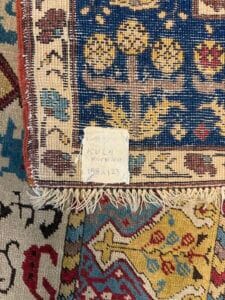 Azerbaijan Tappeti Milano Un primo piano di un angolo di tappeto intrecciato mostra motivi intricati e una piccola etichetta scritta a mano che recita "KULA ANTIK 200 198 x 123". Sotto di esso è visibile anche parte di un altro tappeto colorato con disegni geometrici.