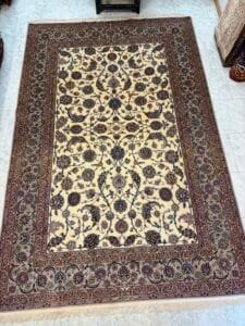 Azerbaijan Tappeti Milano Un tappeto rettangolare ornato con un intricato motivo floreale in beige, marrone e colori scuri, posto su un pavimento piastrellato chiaro. Nell'angolo in alto a destra è presente un logo colorato con una foglia stilizzata.