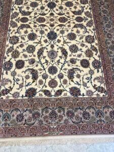 Azerbaijan Tappeti Milano Tappeto decorativo in stile persiano con intricati motivi floreali nei toni del blu, del bordeaux e del beige. Il logo di una farfalla colorata si trova nell'angolo superiore destro dell'immagine. Il tappeto è posizionato su un pavimento chiaro.