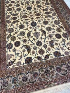 Azerbaijan Tappeti Milano Tappeto persiano color crema con intricati motivi floreali e di viti in blu scuro, rosso e beige, delimitati da un motivo dettagliato. Nell'angolo in alto a destra compare il logo di una farfalla colorata.