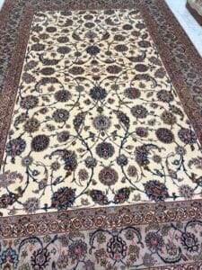 Azerbaijan Tappeti Milano Un grande tappeto ornato in stile persiano con sfondo crema, intricati motivi floreali e di viti in marrone, blu e bordeaux e un dettagliato bordo decorativo, steso su un pavimento chiaro. Il logo di una foglia multicolore si trova in alto a destra.