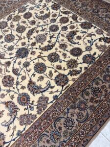 Azerbaijan Tappeti Milano Tappeto beige a fantasia con intricati disegni floreali e di viti nei toni del blu e del marrone, delimitati da un dettagliato bordo geometrico. Nell'angolo in alto a destra compare un logo a forma di foglia colorata.