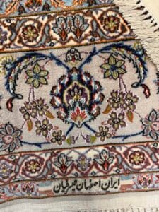 Azerbaijan Tappeti Milano Primo piano di un dettagliato tappeto persiano con intricati motivi floreali e colori vivaci. In basso, la scrittura persiana recita "ایران اصفهان قیمتیان", indicando che proviene da Isfahan, Iran. Nell'angolo sono visibili alcune frange.