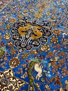 Azerbaijan Tappeti Milano Un tappeto persiano dettagliato con motivi intricati in blu, marrone e oro, con motivi floreali e animali tra cui uccelli. Un logo multicolore è nell'angolo in alto a destra.
