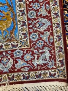 Azerbaijan Tappeti Milano Primo piano di un angolo di tappeto decorativo con intricati motivi floreali e di uccelli in rosso, blu e beige. Un disegno di farfalla colorata è in alto a destra. Una frangia bianca è in basso.