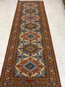 Azerbaijan Tappeti Milano Un lungo tappeto rettangolare con disegni geometrici in blu, rosso, marrone e beige, steso su un pavimento di marmo chiaro. Un logo colorato e stilizzato appare nell'angolo in alto a destra.