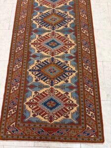 Azerbaijan Tappeti Milano Un lungo tappeto rettangolare con intricati disegni geometrici nei colori rosso, blu, oro e crema. Il tappeto è posizionato su un pavimento piastrellato chiaro, con un logo colorato nell'angolo in alto a destra.