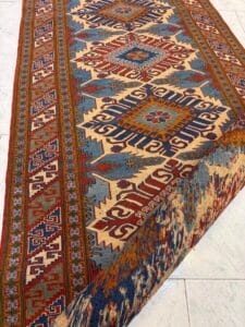 Azerbaijan Tappeti Milano Un tappeto colorato e intricato con disegni geometrici in blu, rosso, giallo e beige, parzialmente piegato in un angolo, esposto su un pavimento piastrellato chiaro.