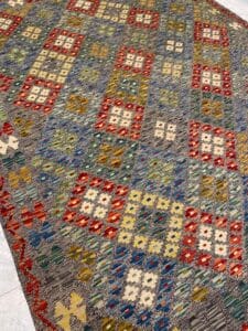 Azerbaijan Tappeti Milano Un tappeto colorato con motivi geometrici in rosso, giallo, blu e verde su sfondo grigio, caratterizzato da un piccolo motivo a foglie multicolori in un angolo.