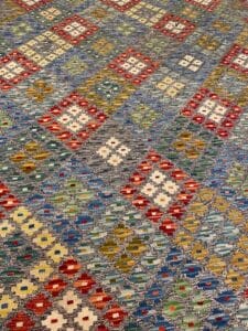 Azerbaijan Tappeti Milano Un tappeto colorato con motivi geometrici a forma di rombi rossi, bianchi, gialli e verdi su uno sfondo grigio. Un piccolo logo multicolore si trova nell'angolo in alto a destra.
