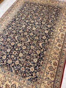 Azerbaijan Tappeti Milano Un tappeto con motivi floreali e geometrici intricati nei toni del blu e del beige. Un piccolo logo colorato è posizionato nell'angolo in alto a destra.