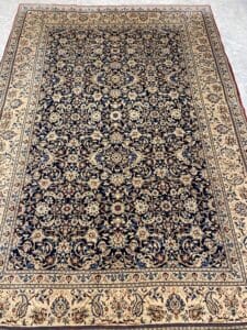 Azerbaijan Tappeti Milano Un tappeto persiano dettagliato con intricati motivi floreali nei toni del blu navy, crema e rosso su un pavimento piastrellato. Un piccolo logo a forma di foglia colorata si trova nell'angolo in alto a destra.
