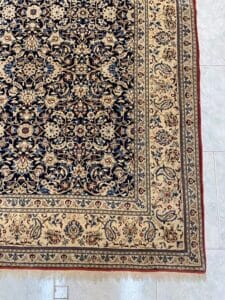 Azerbaijan Tappeti Milano Primo piano di un tappeto con motivi floreali intricati in tonalità blu scuro, beige e rosso. L'angolo mostra un logo a forma di stella color arcobaleno su uno sfondo di pavimento piastrellato.