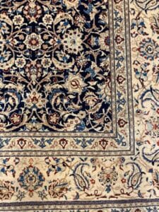 Azerbaijan Tappeti Milano Primo piano di un tappeto orientale con motivi floreali in blu, crema e marrone. Un piccolo logo a forma di foglia colorata si trova nell'angolo in alto a destra.