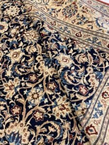Azerbaijan Tappeti Milano Un tappeto persiano dettagliato con intricati motivi floreali in blu, rosso e beige, con un logo colorato parzialmente visibile in un angolo.