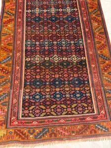 Azerbaijan Tappeti Milano Tappeto tradizionale con intricati motivi geometrici in rosso, blu, giallo e arancione. I bordi sono colorati e nell'angolo in alto a destra compare il logo di una farfalla multicolore. Il tappeto è appoggiato su una superficie chiara.