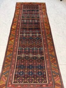 Azerbaijan Tappeti Milano Un lungo tappeto rettangolare in stile persiano con intricati motivi geometrici nei toni del rosso, del blu e del beige è steso su un pavimento piastrellato chiaro. Un logo colorato a forma di foglia con una "A" si trova nell'angolo in alto a destra.