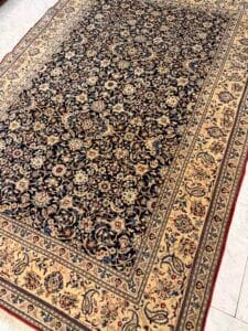 Azerbaijan Tappeti Milano Tappeto in stile persiano con intricati disegni floreali in colori scuri e chiari, delimitati da una fascia floreale marrone. Nell'angolo superiore destro dell'immagine compare un logo astratto multicolore.