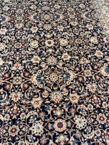 Azerbaijan Tappeti Milano Tappeto dettagliato e ornato in stile persiano con intricati motivi floreali in beige, blu e rosso. Nell'angolo superiore destro, un logo colorato a forma di foglia con sezioni rosse, verdi, gialle e arancioni si sovrappone al tappeto.