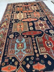 Azerbaijan Tappeti Milano Tappeto orientale colorato e dai motivi intricati, con forme geometriche nei toni del rosso, del blu, del verde e dell'arancione, esposto su un pavimento chiaro. Nell'angolo in alto a destra è visibile il logo di una foglia multicolore.