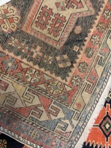 Azerbaijan Tappeti Milano Primo piano di un tappeto a motivi intrecciati con forme geometriche nei toni del rosso, del blu, del beige e del nero; nell'angolo in alto a destra compare il logo di una foglia colorata con la parola "Jaly".