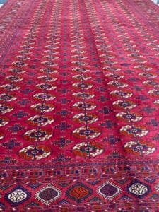 Azerbaijan Tappeti Milano Tappeto rosso con motivi geometrici, caratterizzato da una foglia colorata e dal logo "A" nell'angolo in alto a destra.