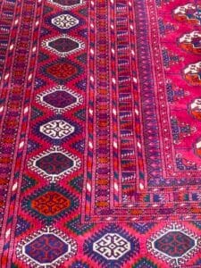 Azerbaijan Tappeti Milano Un vivace tappeto con motivo geometrico nei toni del rosso, viola e verde, con un logo a forma di foglia colorata nell'angolo.