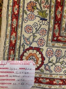 Azerbaijan Tappeti Milano Primo piano di un tappeto decorato con motivi floreali e un'etichetta in italiano che ne descrive l'origine, la composizione, le dimensioni e il prezzo.
