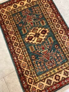Azerbaijan Tappeti Milano Tappeto rettangolare con motivi geometrici nei toni del rosso, verde e beige su pavimento piastrellato.