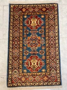 Azerbaijan Tappeti Milano Un tappeto orientale rettangolare con centro blu, motivi rossi e beige e bordo geometrico.