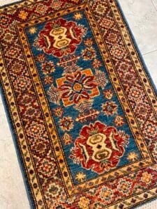 Azerbaijan Tappeti Milano Un colorato tappeto orientale con motivi geometrici rossi, dorati e blu su un pavimento piastrellato.