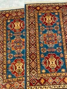 Azerbaijan Tappeti Milano Due tappeti colorati con motivi geometrici blu, rossi e gialli su un pavimento piastrellato.