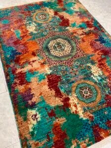 Azerbaijan Tappeti Milano Un tappeto multicolore con motivi geometrici e ornamentali nelle tonalità del verde acqua, ruggine, beige e arancione su un pavimento piastrellato chiaro.