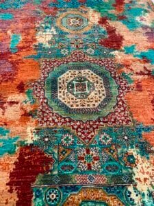 Azerbaijan Tappeti Milano Un tappeto orientale multicolore con motivi geometrici e floreali nei toni del verde acqua, ruggine e crema, caratterizzato da un logo decorativo con una foglia colorata nell'angolo in alto a destra.